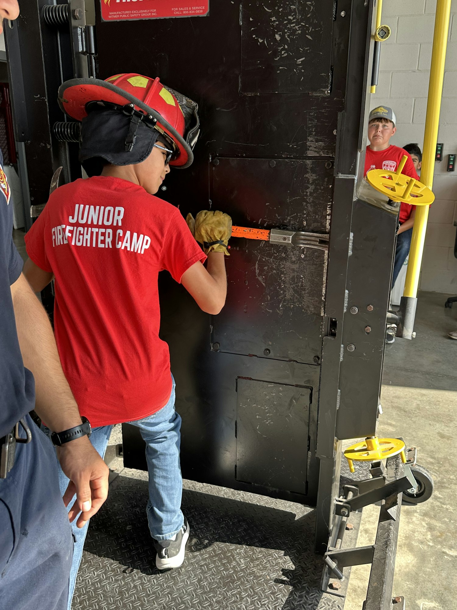 A child in a red "Junior Firefighter Camp" shirt uses a tool on a black training apparatus, supervised by adults.