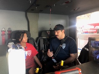 A child and an adult in uniform are sitting inside a rescue vehicle, having a conversation.