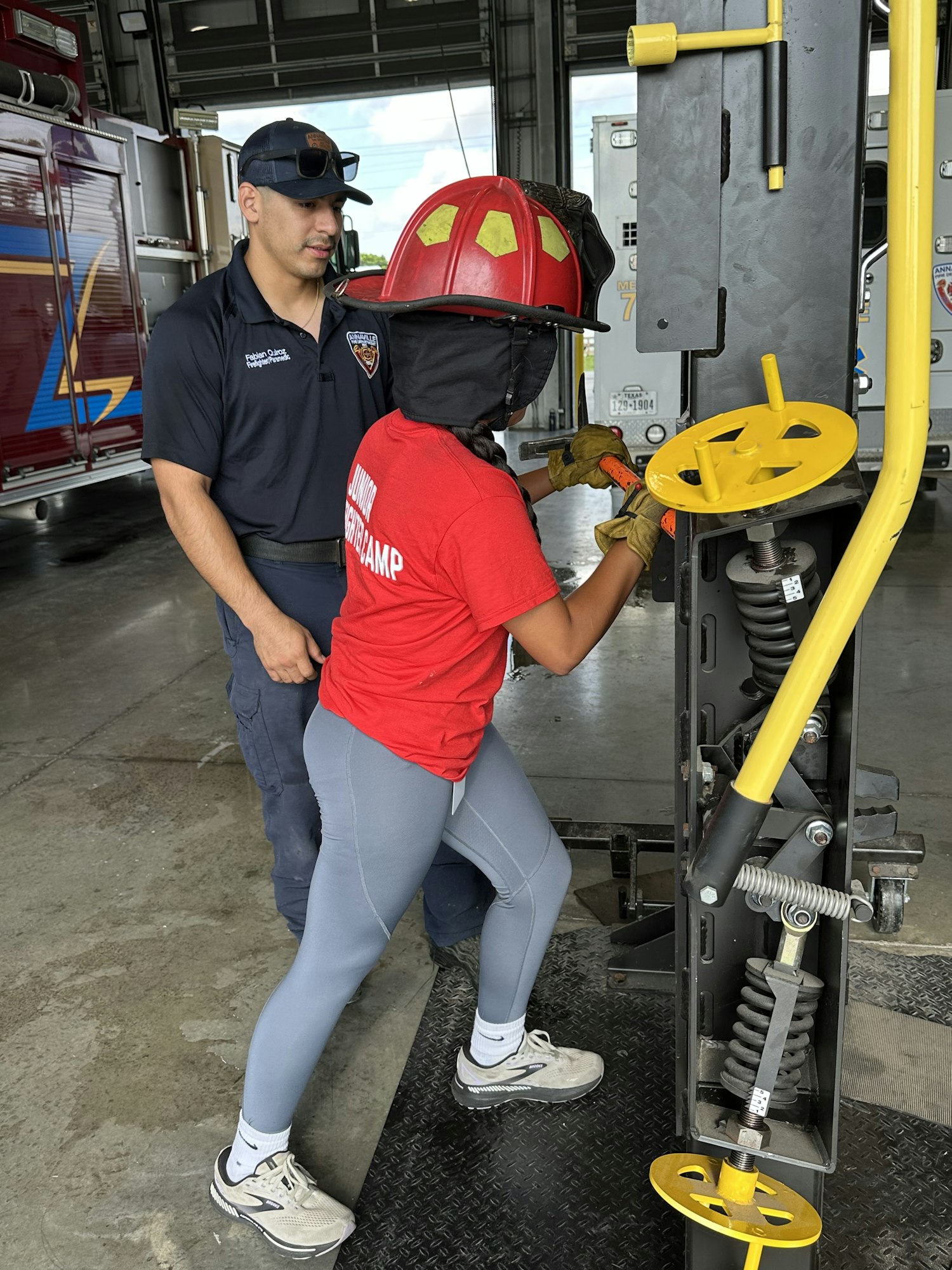 A person in a red shirt and safety gear operates a tool with firefighter supervision.
