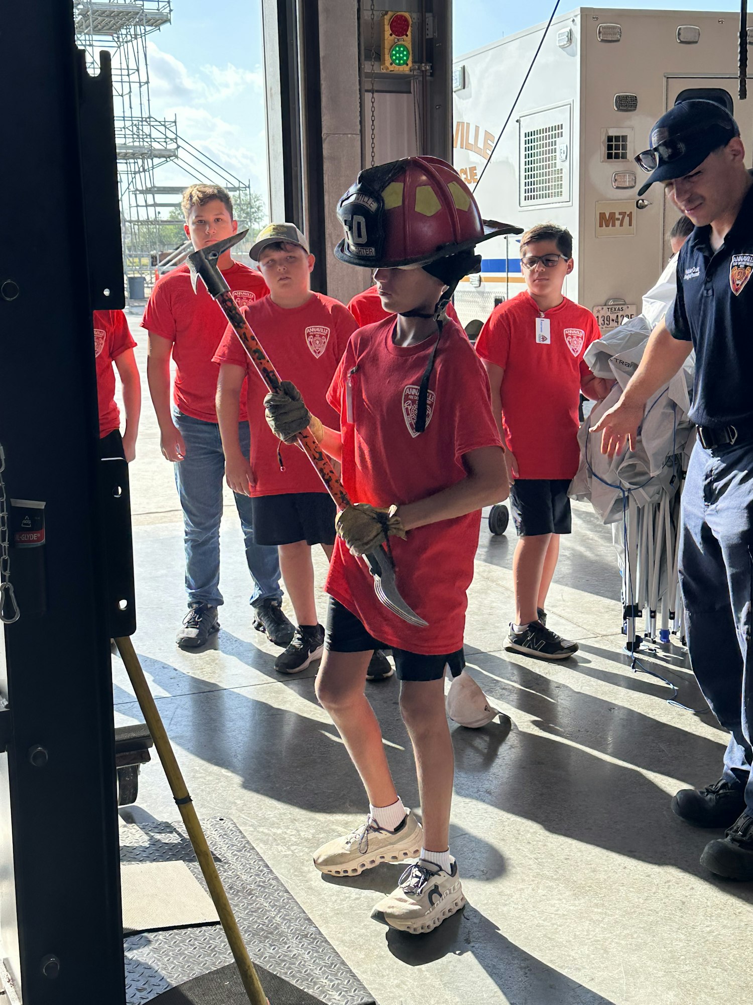 A group of people in red shirts; one is wearing a firefighter helmet and holding an axe-like tool.