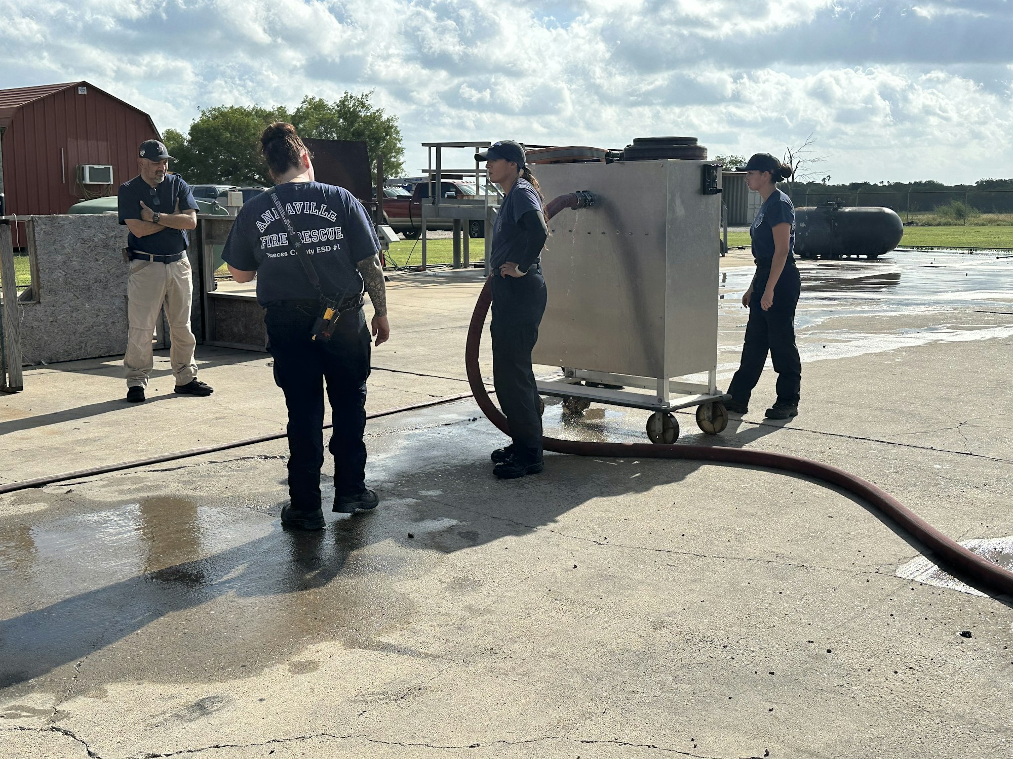 People in fire rescue gear, standing outside by equipment and a hose on a sunny day.