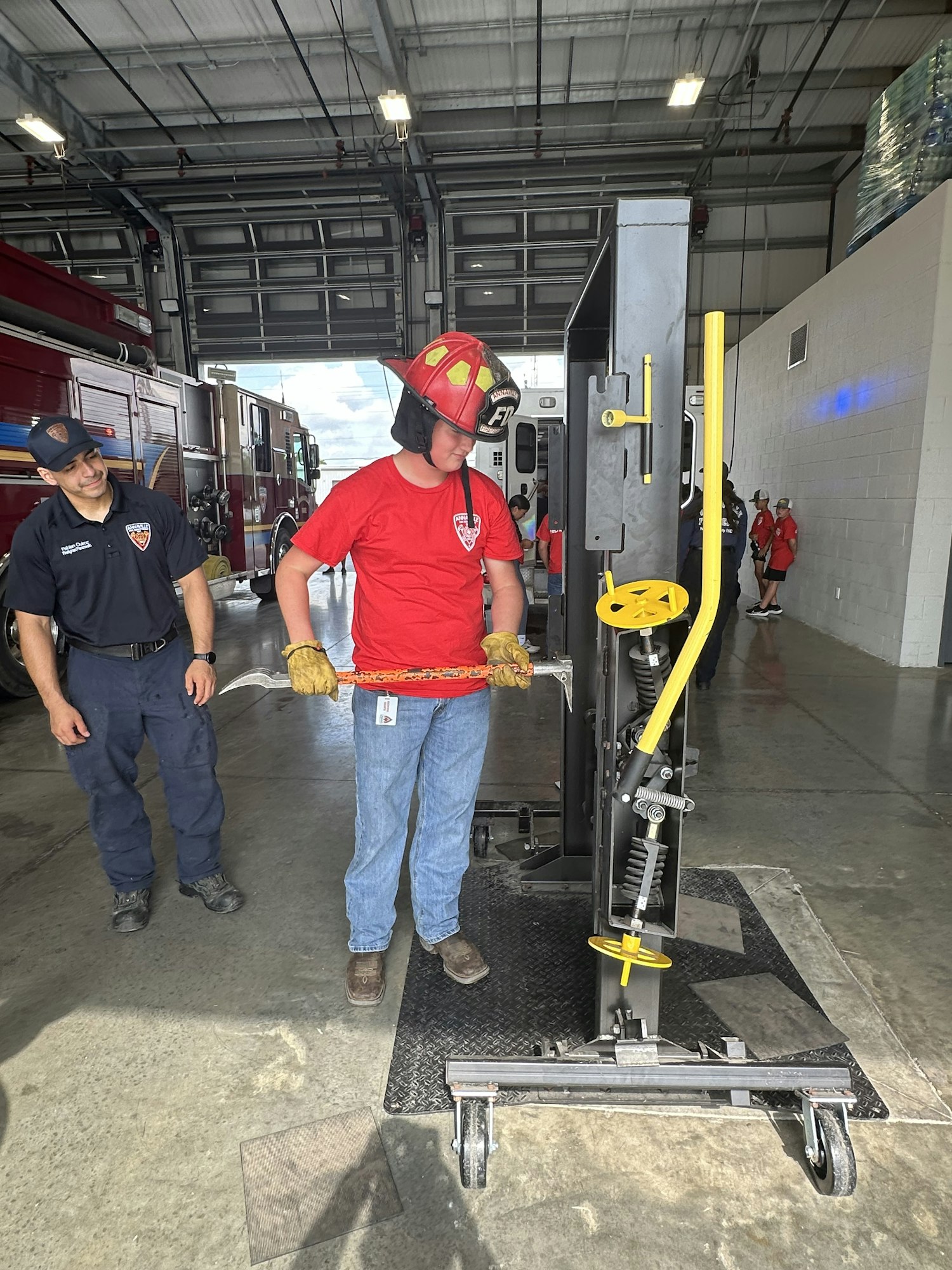 Two individuals in a fire station with firefighting equipment; one wears a helmet and holds a tool, the other observes.