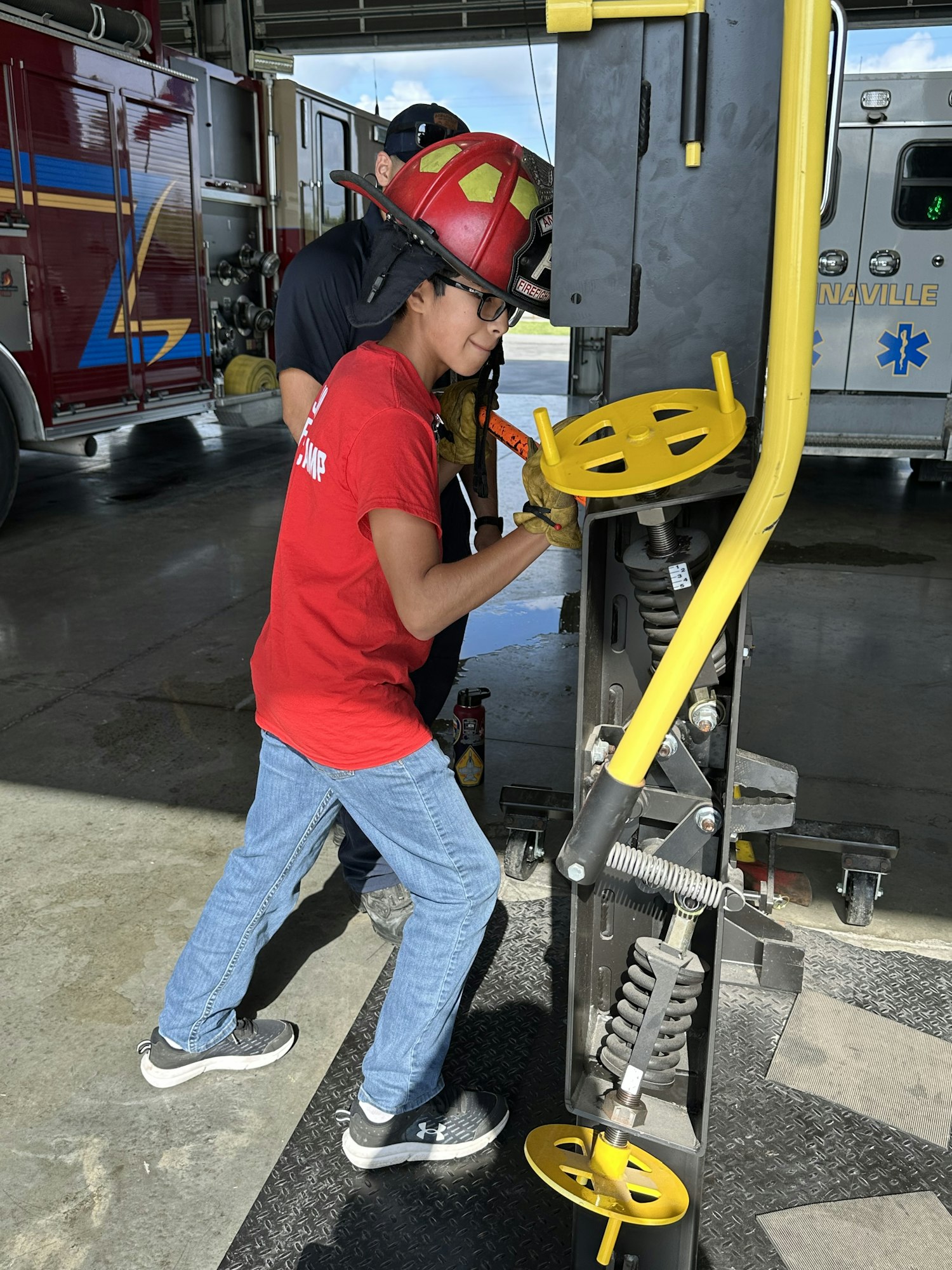 Person in a red shirt and helmet using a tool at a fire station. A fire truck and emergency equipment are visible.