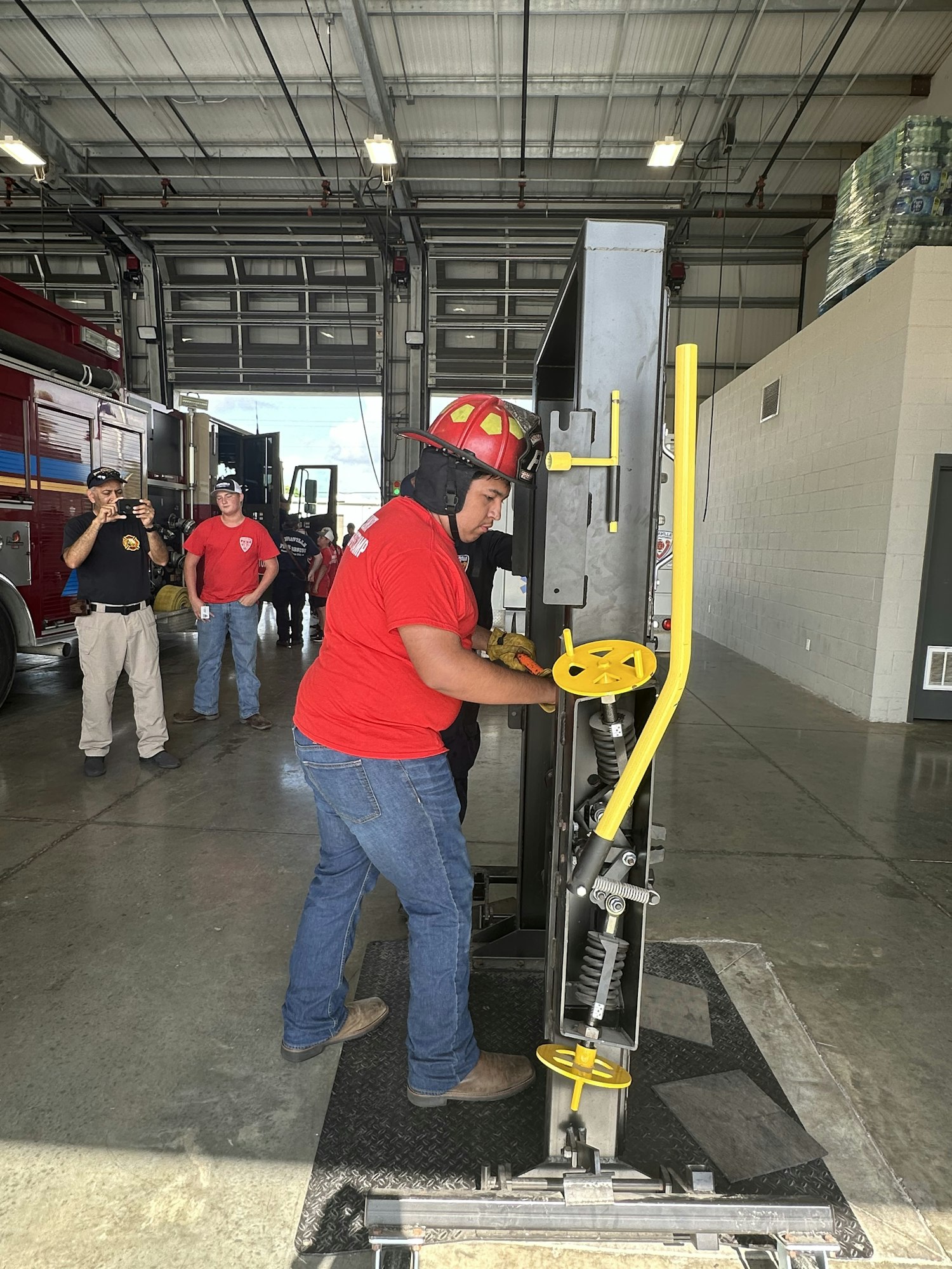 A person in a firefighter helmet uses a training device in a fire station with others watching.