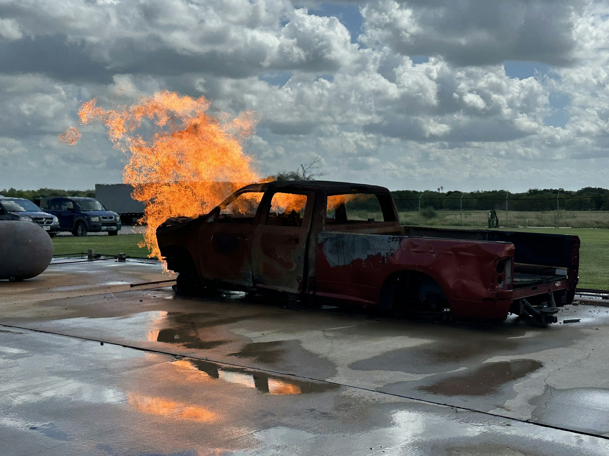 A burnt-out truck with flames coming from the front, parked outdoors on a wet surface.
