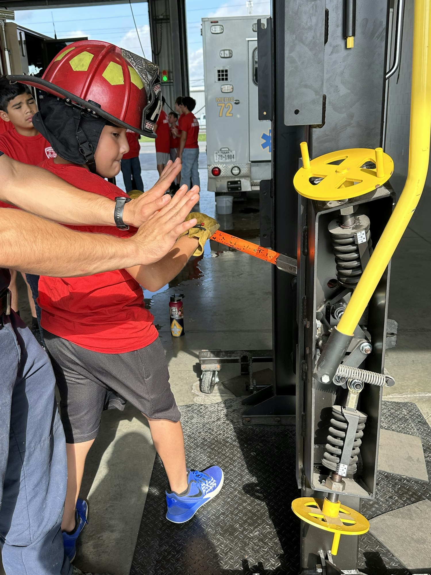 A child wearing a firefighter helmet uses a tool with the help of an adult, while others look on in the background.