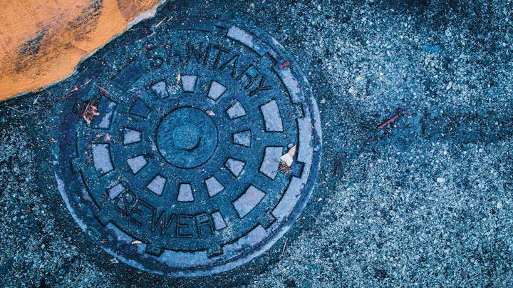 A manhole cover labeled "SANITARY SEWER" on a textured asphalt surface with orange paint and scattered leaves.