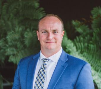 A person in a blue suit and plaid tie standing in front of a green leafy background.