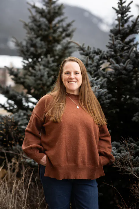 A smiling person in a brown sweater stands outdoors in front of evergreen trees, with a mountainous background.