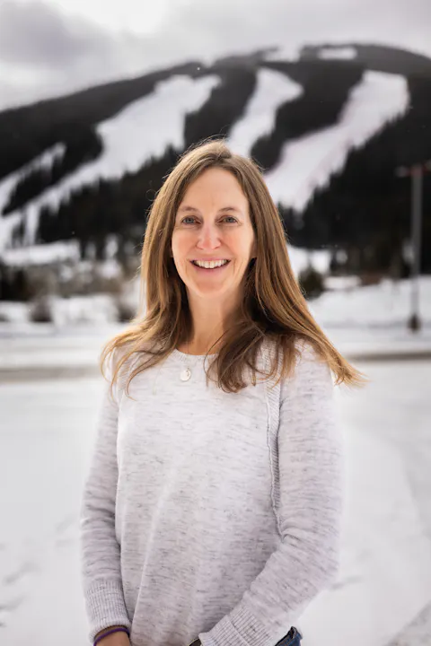 A woman smiles in a snowy landscape with a mountain backdrop and ski slopes visible. The scene is bright and wintery.