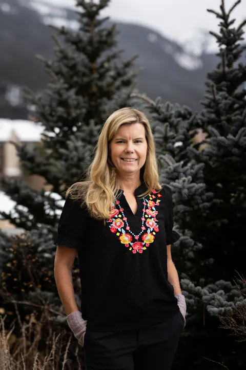 A woman stands outdoors in front of evergreen trees, wearing a black top with colorful floral embroidery.