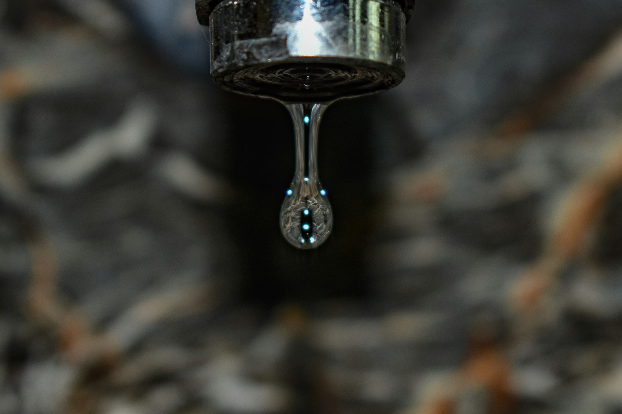Close-up of a drop of water hanging from a faucet tip, with a blurred background.