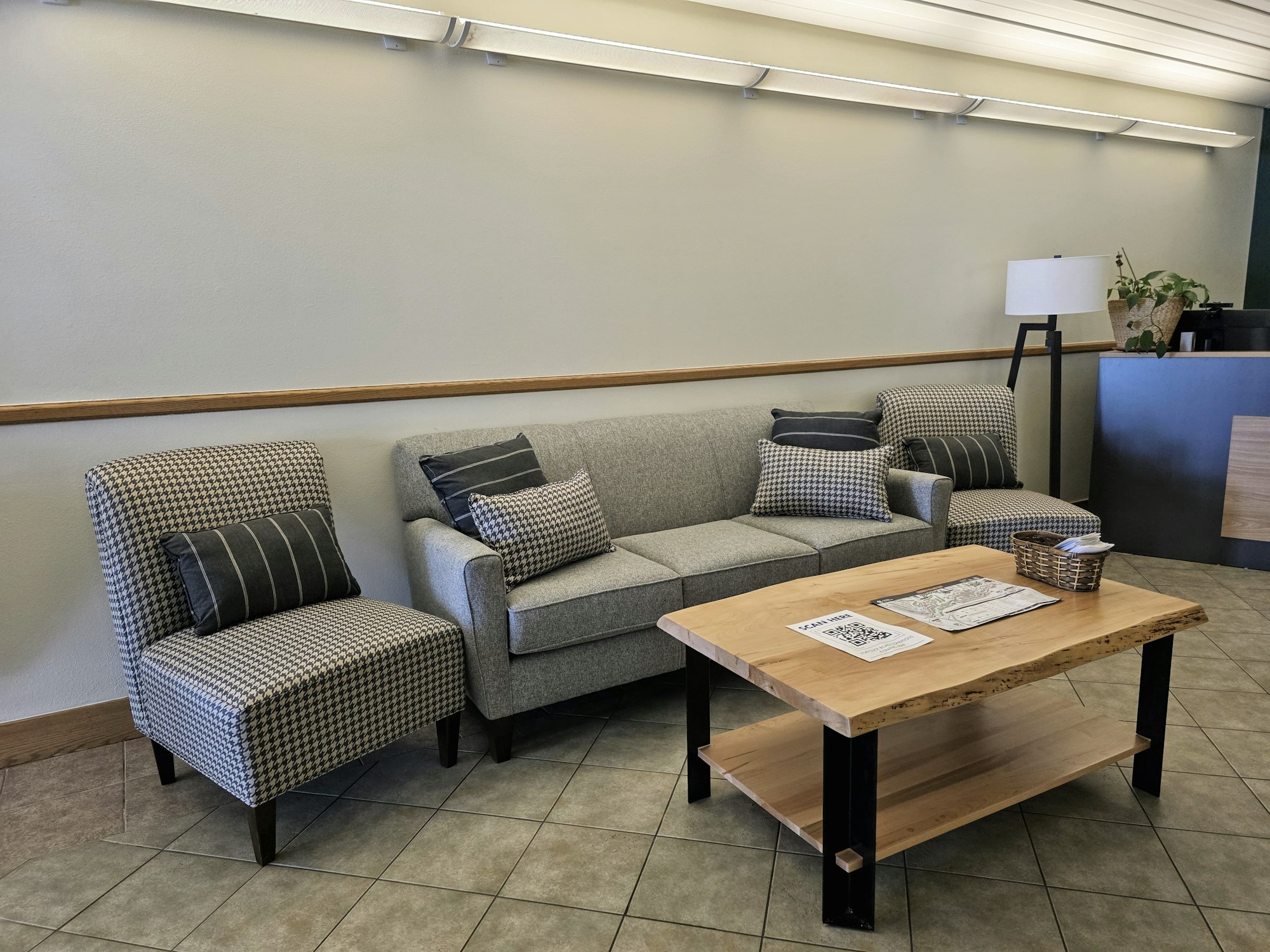 Gray couch and chairs with pillows in a waiting room, a wooden coffee table with papers and a basket, tile floor, lamp, and plants.