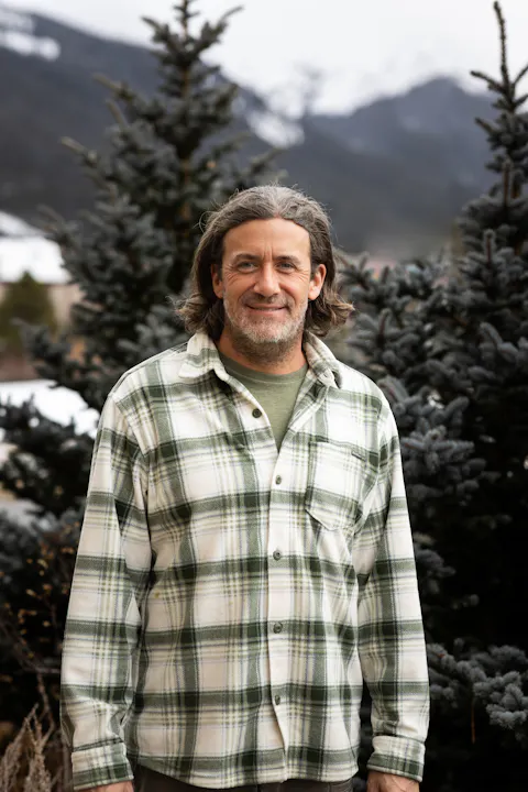 A man with long hair, wearing a plaid flannel shirt, stands outdoors amidst trees and mountains under a cloudy sky.
