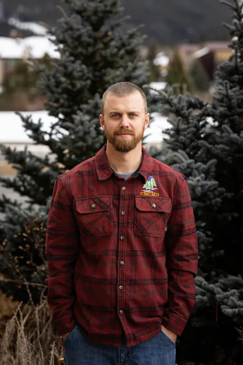 A man in a red plaid shirt stands outdoors with evergreen trees in the background, presumably in a winter setting.