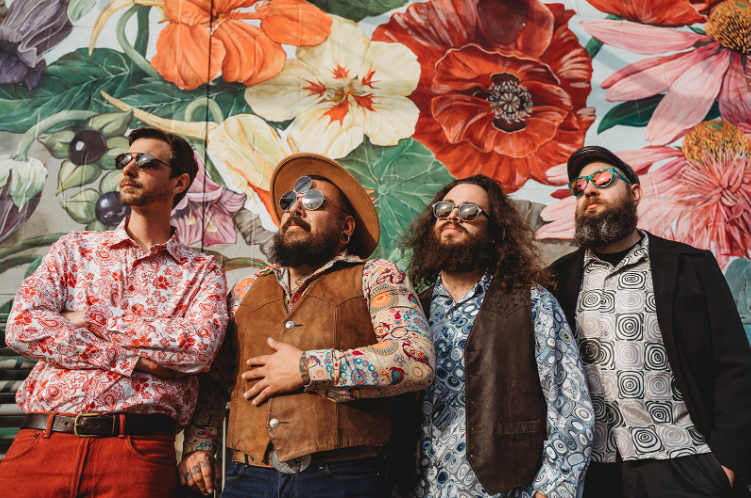 Four men in vintage outfits and sunglasses pose confidently in front of a colorful floral mural.
