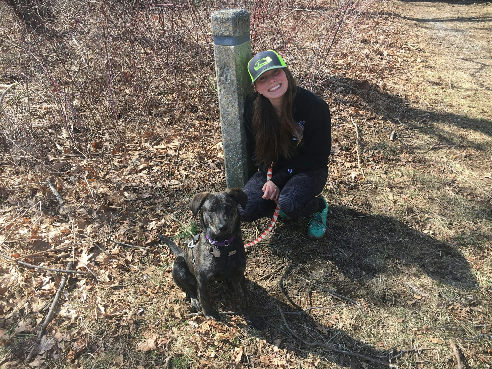 A woman and a dog posing next to a trail marker outdoors.