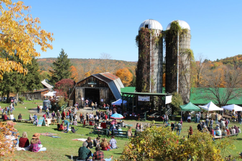 Outdoor event at a farm with barns, silos, people relaxing, and tents on a sunny day.