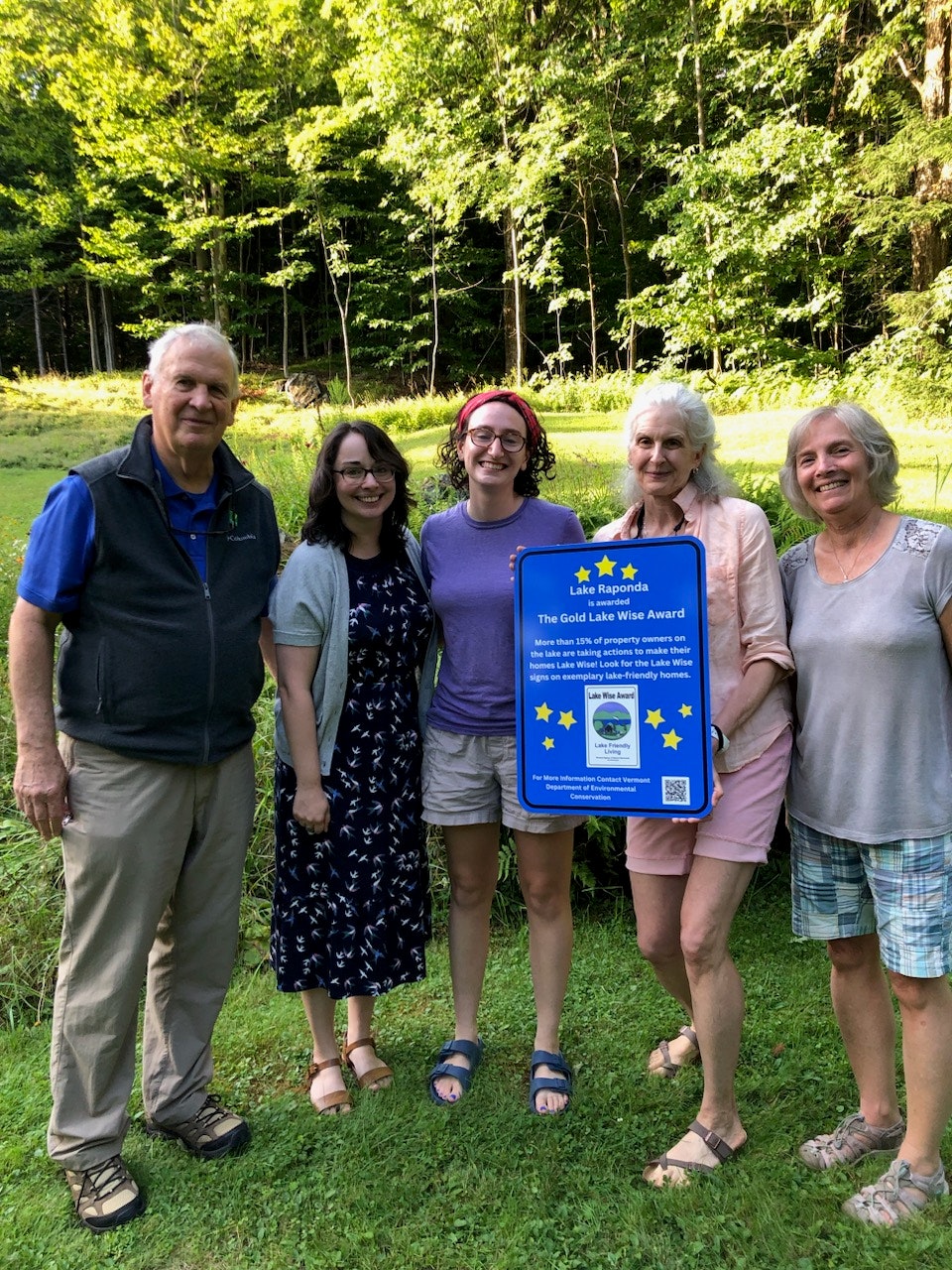Five people standing outdoors, holding a "Lake Wise Award" sign in front of a forest background.