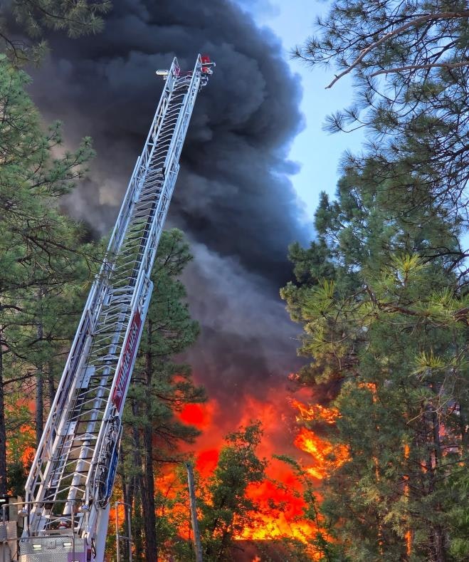 A fire truck's ladder reaches toward thick smoke and flames, surrounded by trees, indicating an ongoing fire emergency.