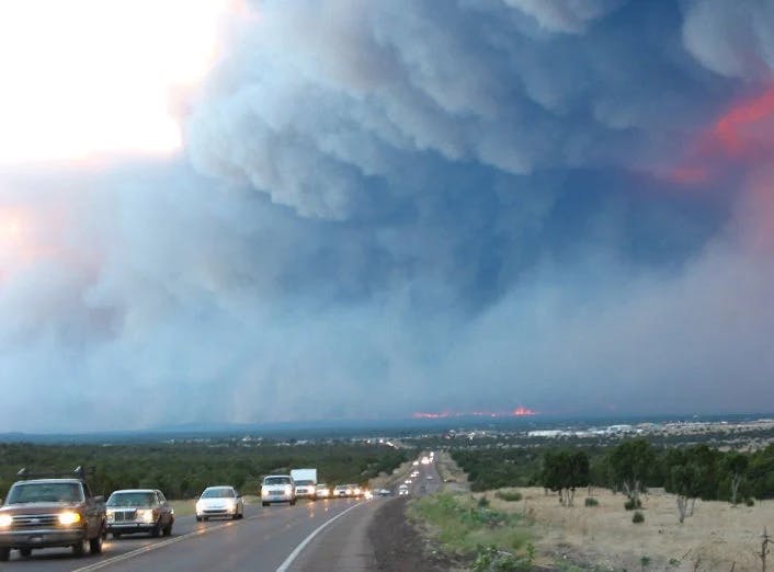 Cars on a road drive away from a massive smoke plume rising from a distant wildfire.