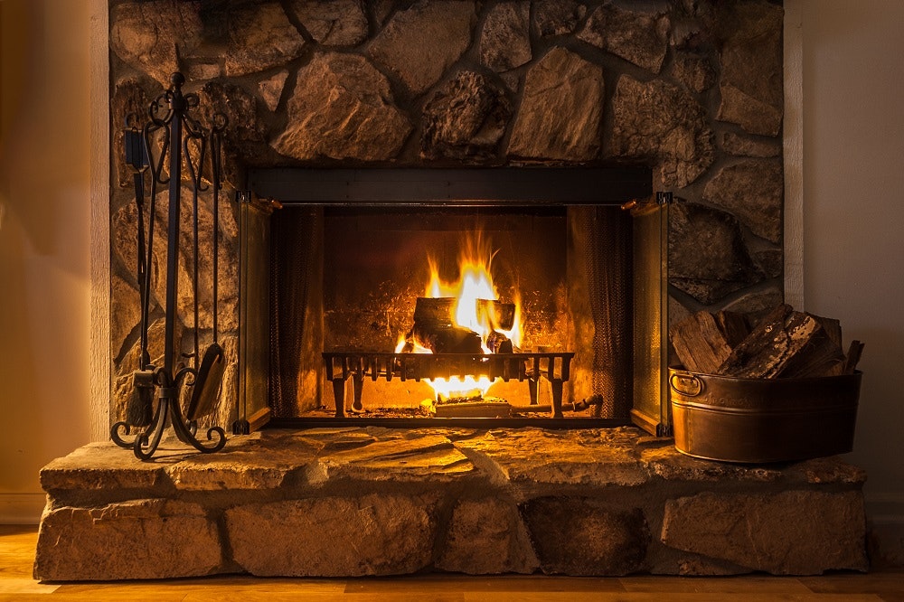 A lit stone fireplace with logs burning, fireplace tools on the left, and a basket of firewood on the right.
