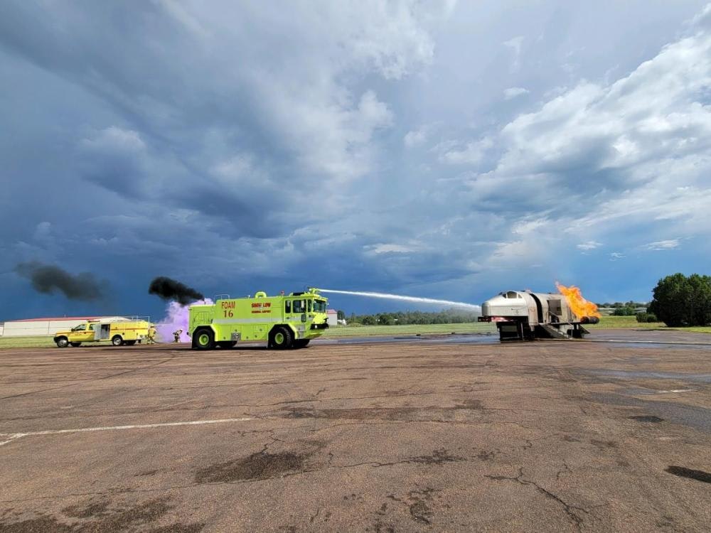 Firefighting drill with trucks using hoses to put out a controlled fire on an aircraft fuselage.