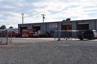 A parking area with a fire truck, vehicles, and a building in the background, behind a chain-link fence.