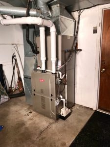 A residential furnace system installed in a basement with surrounding pipes and a door to the side.