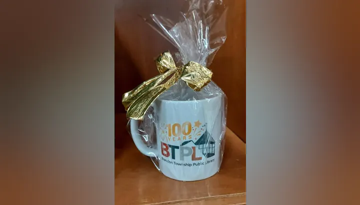A festive mug celebrating 100 years of the Brandon Township Public Library, wrapped in clear plastic with a gold bow.