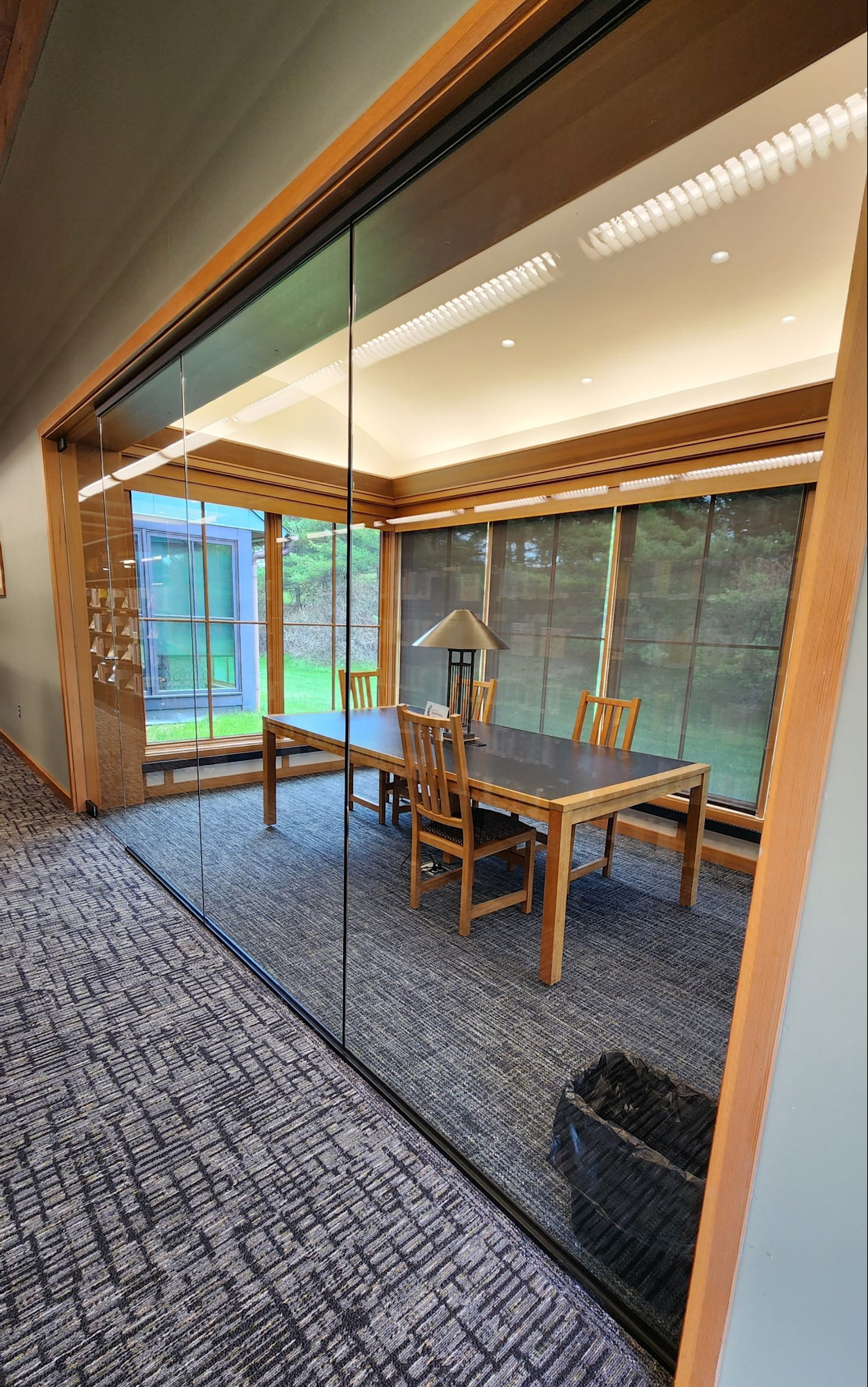 A modern conference room with a wooden table, chairs, a lamp, and large windows overlooking greenery.
