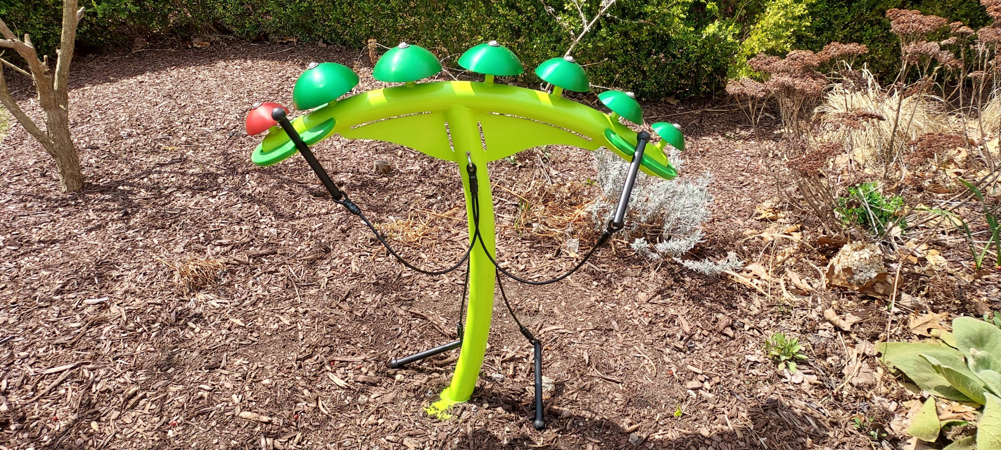 Outdoor musical instrument with green and red concave shapes, resembling bells or saucers, set in a garden area.