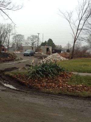 A gloomy day with a dirt road, leaf pile, green plants, a car, and construction materials in the background.