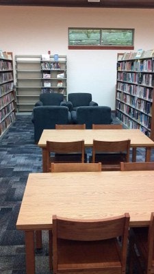 A library interior with bookshelves, tables, chairs, and a seating area with armchairs.