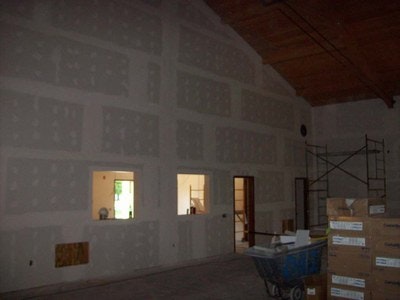 An interior under construction with drywall installation, a scaffold, a wheelbarrow, and building materials.