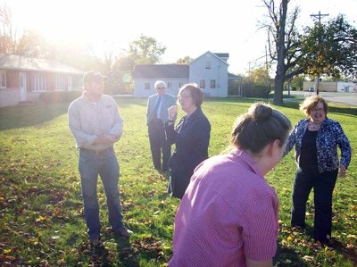 A group of people standing outdoors on a sunny day with fallen leaves on the ground, houses in the background.