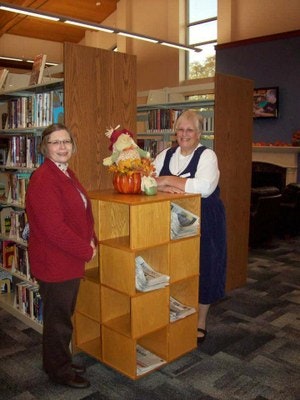 Two women in a library with books and a decorative arrangement on a wooden structure.