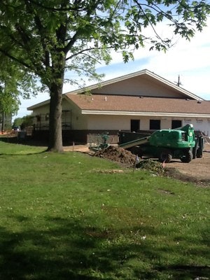 A building under construction with a green portable toilet and construction equipment surrounded by trees.