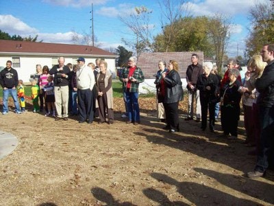 Group of people gathered outdoors, possibly for an event, with trees and a building in the background.