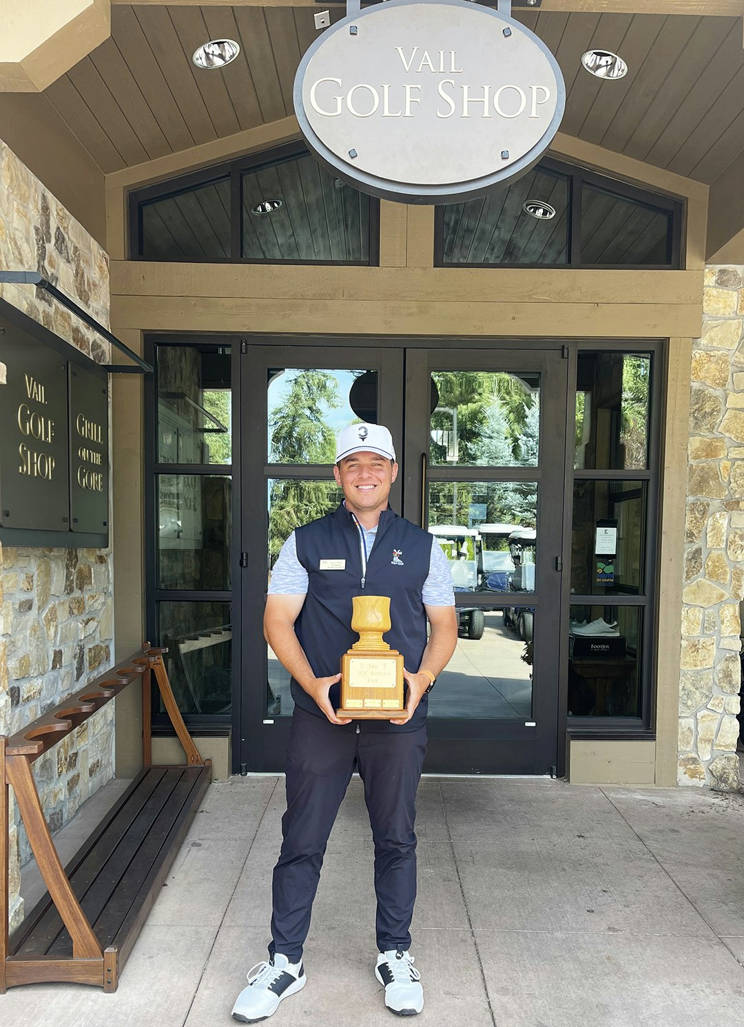 A person stands outside a golf shop, happily holding a wooden trophy.