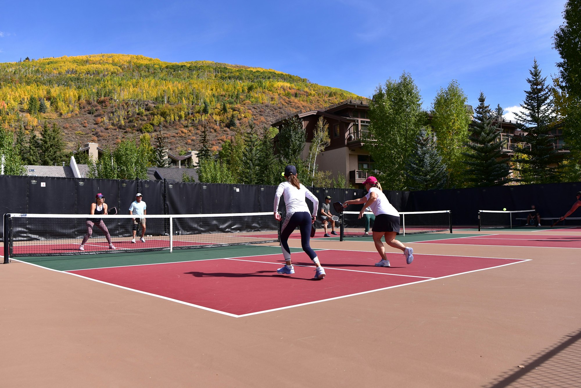 People are playing tennis on an outdoor court with colorful trees in the background on a sunny day.