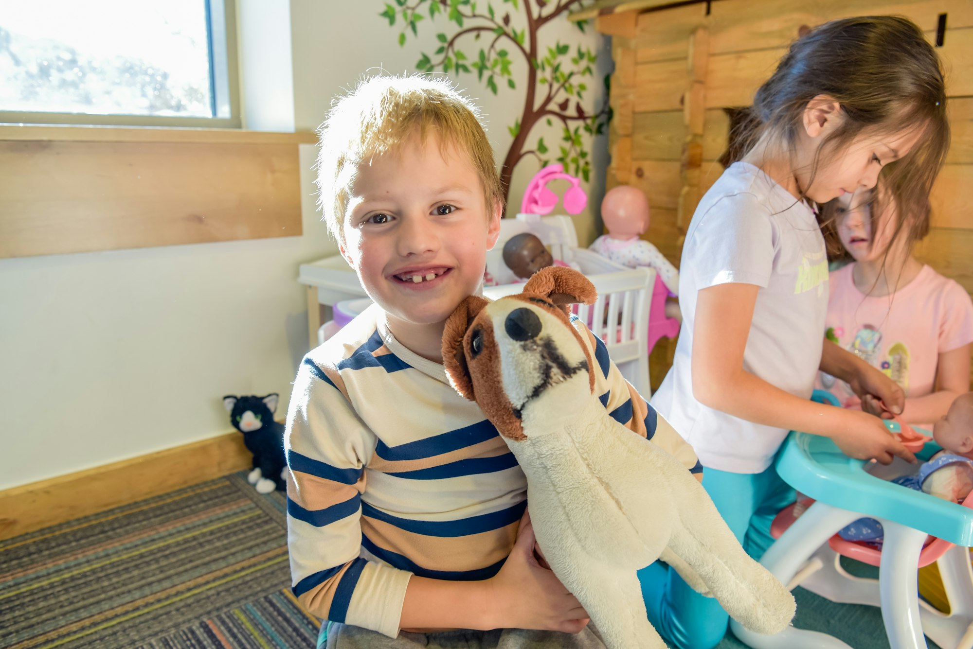 A smiling child holding a plush dog, with other children playing in a room filled with toys.