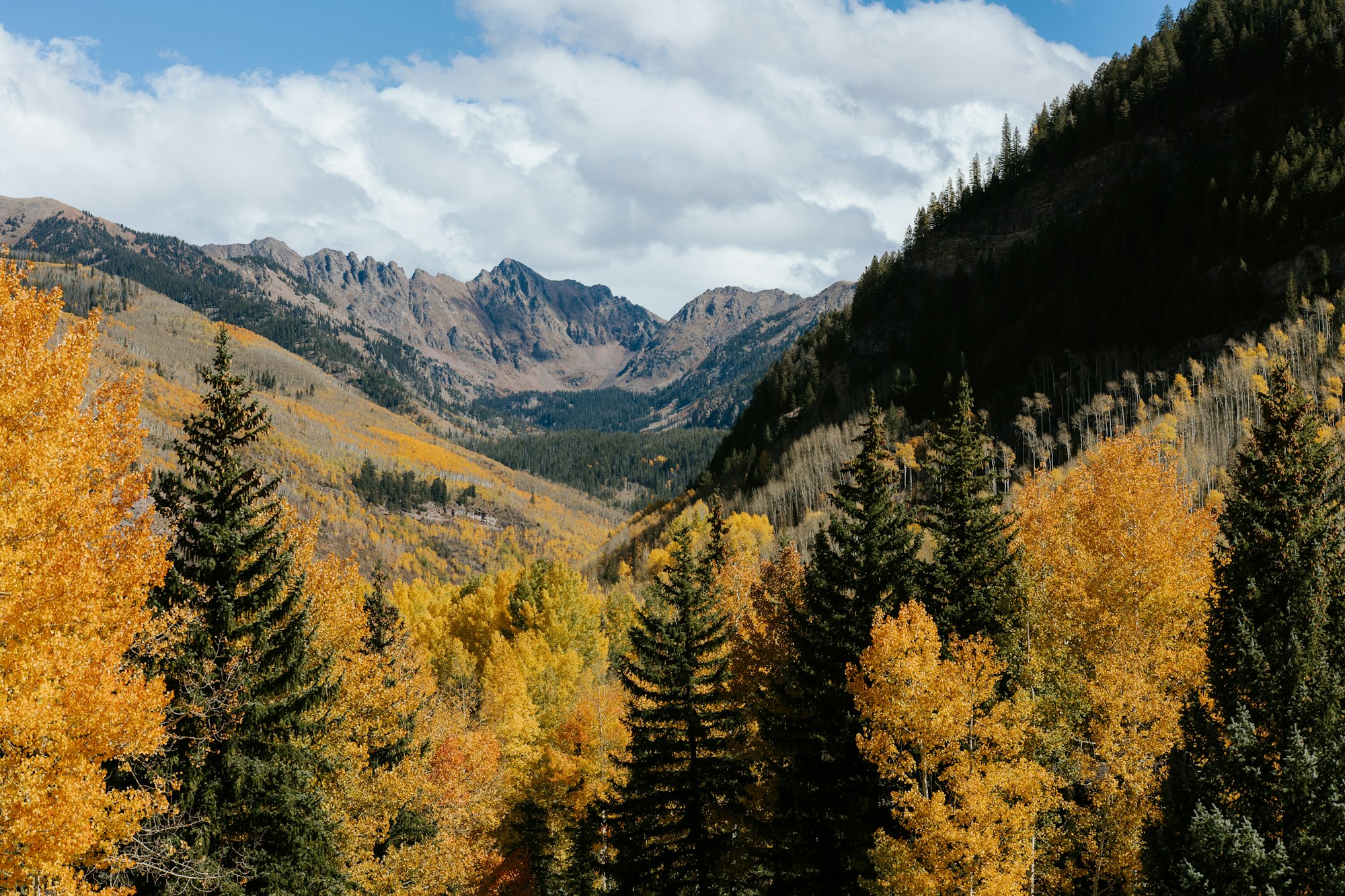 A scenic mountain landscape featuring vibrant fall foliage, with towering peaks and a blue sky dotted with clouds.