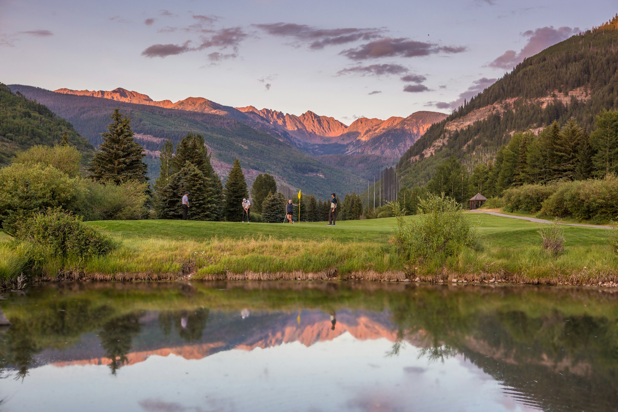 Sunset-lit mountains, golfers, reflecting pond, and lush greenery.