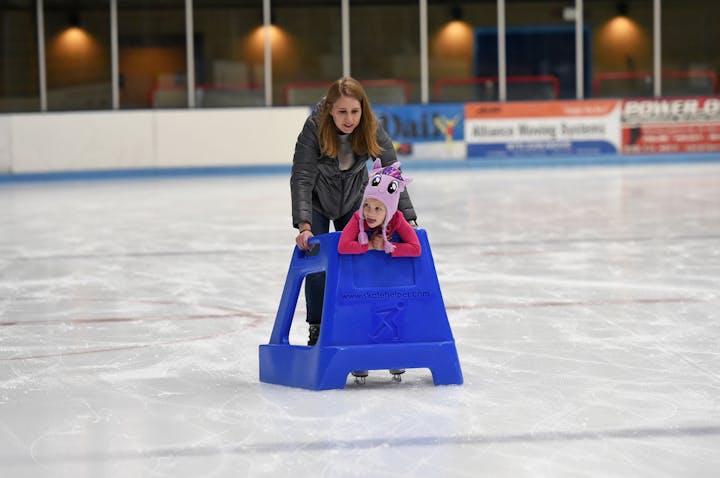 A woman assists a child using a training aid on an ice skating rink.