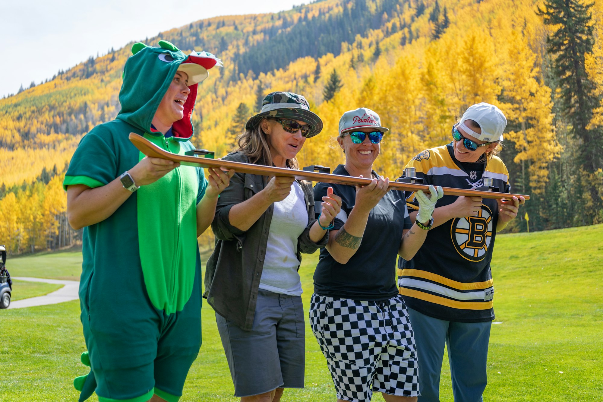 Four people in festive attire holding a ski and smiling outdoors with autumn trees in the background.