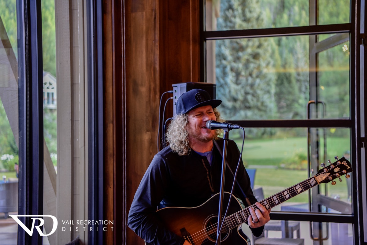 A musician playing guitar and singing into a microphone indoors with a natural backdrop visible through windows.
