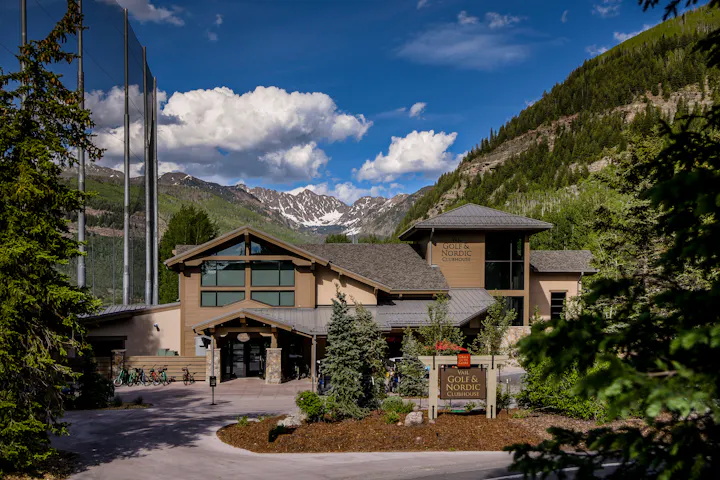 The image shows the Vail Golf & Nordic Clubhouse, set against a backdrop of mountains and blue skies. Trees and bicycles are visible.