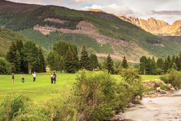 A scenic golf course with players, a river, trees, and mountains under a partly cloudy sky.