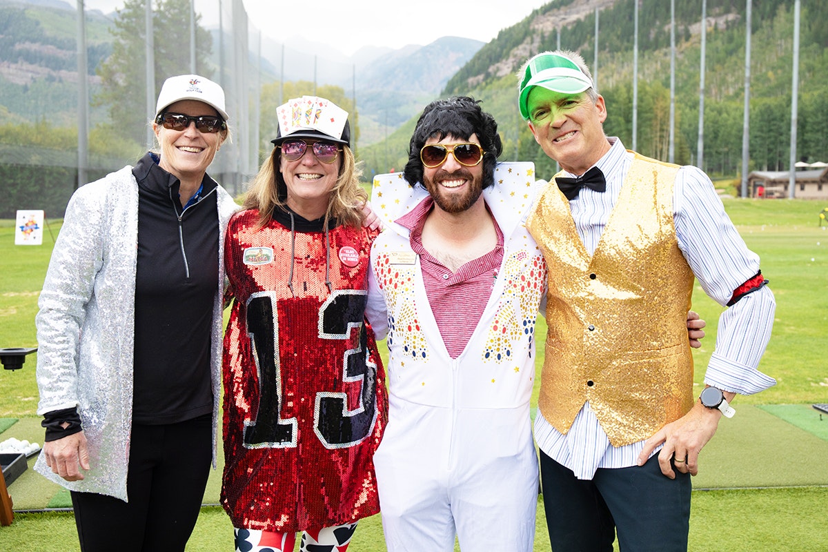 Four people in costumes, with a sequined sports jersey, Elvis outfit, and festive attire, posing outdoors.
